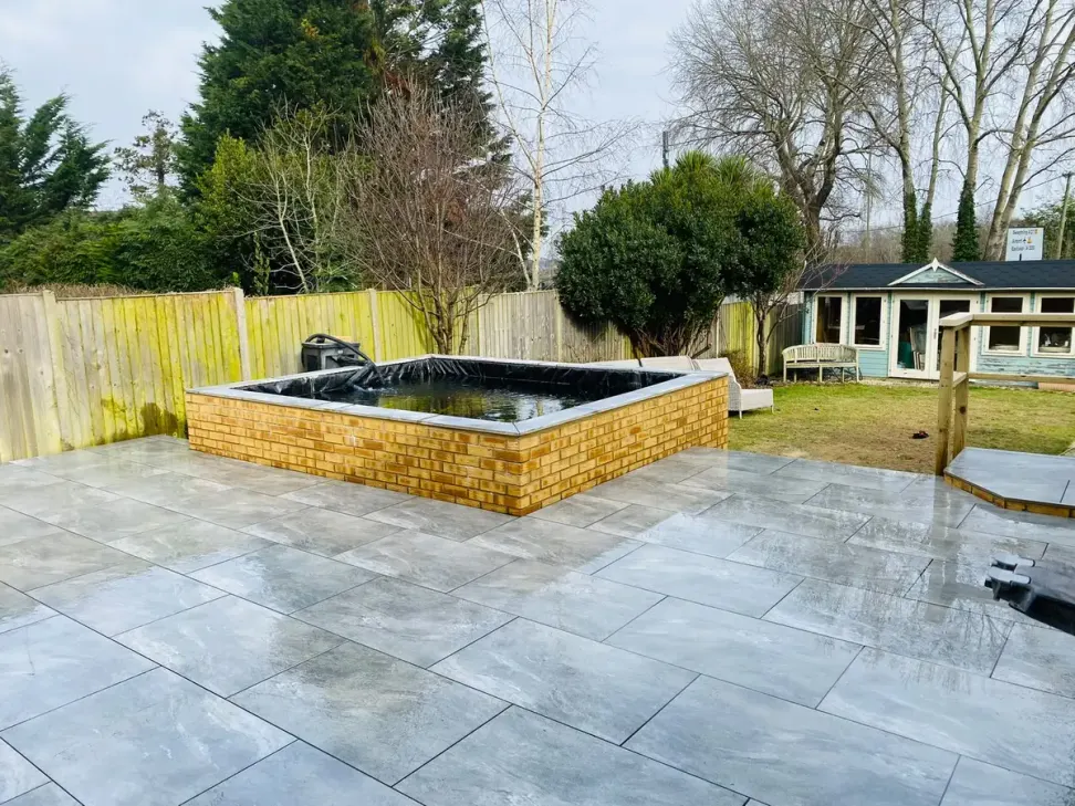 Wide grey porcelain patio with brick raised pond and summerhouse behind